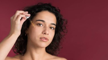 Genç beyaz bir kadın yüzüne bir bakım ürünü uyguluyor, kameraya bakıyor.