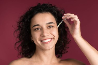 Genç beyaz bir kadın yüzüne bakım ürünü uyguluyor, kameraya bakıyor.