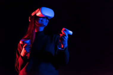 VR oyunlarıyla ilgilenen genç bir kadın VR gözlük ve kumanda kullanıyor. Kırmızı ve mavi aydınlatma