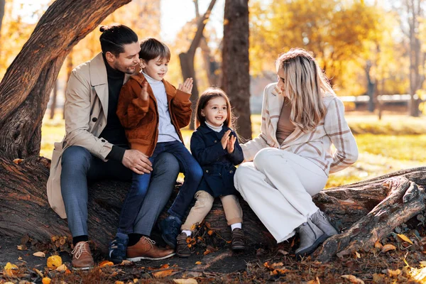 stock image Happy family in an autumn park. Mother, father, son and daughter are sitting on a tree trunk, yellowed trees around