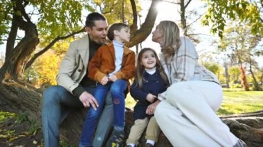 Sonbahar parkında mutlu bir aile. Anne, baba, oğul ve kız ağaç gövdesinde oturuyor, anne oğlunu öpüyor, etrafta sararmış ağaçlar var. Yavaş çekim