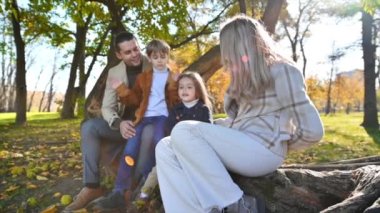 Sonbahar parkında mutlu bir aile. Anne, baba, oğul ve kız bir ağaç gövdesinde oturuyor, etrafta sarı ağaçlar var. Yavaş çekim