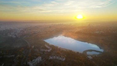 Chisinau 'nun güneş doğarken, Moldova' daki hava aracı görüntüsü. Valea Morilor Parkı 'nın gölü ve sararmış ormanı, etrafında birden fazla bina, havada sis.