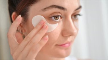 Genç beyaz bir kadın yatakta oturuyor ve yüz masajı yapıyor. Aynaya bakıyor, cilt bakım ürünleri yakınlarda.