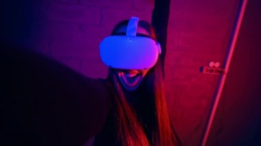 VR oyunlarına ilgi duyan genç bir kadın VR gözlük ve kumanda kullanarak kendini kameraya çekiyor. Kırmızı ve mavi aydınlatma. Yavaş çekim