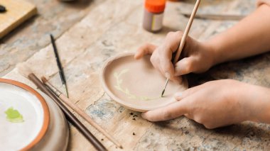 Stüdyoda çalışan bir çömlek ustası. Tabağın altını fırçayla boyamak, etrafta aletler...