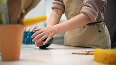 Stüdyoda çalışan bir çömlek ustası. Elleriyle bir parça kil topluyor. Aletler masada. Yavaş çekim