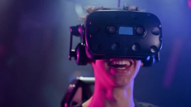 Neşeli, şok olmuş bir çocuk kasklı, kumandalı ve sırt çantalı VR koşu bandında oynuyor. Yakınlarda oynayan diğer insanlar