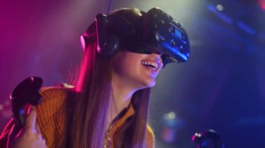 Neşeli, ilgili bir kız VR koşu bandında kask ve kumandayla oynuyor. Yakınlarda oynayan başkaları da var. Yavaş çekim