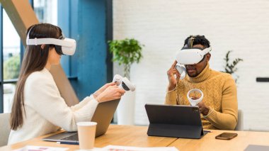 Business conference in VR in an office. A black man and caucasian woman using VR glasses and controllers, papers and gadgets on the table