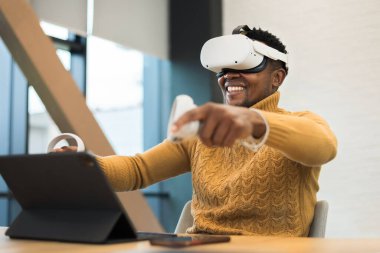 An excited black man exploring virtual reality using VR glasses and controllers in an office, tablet on the table