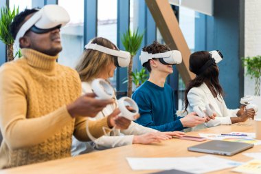 Business conference in VR in an office. Multiracial group of people using VR glasses and controllers, papers and gadgets on the table