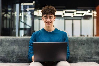 A young smiling man working using a laptop while sitting on a sofa in an office