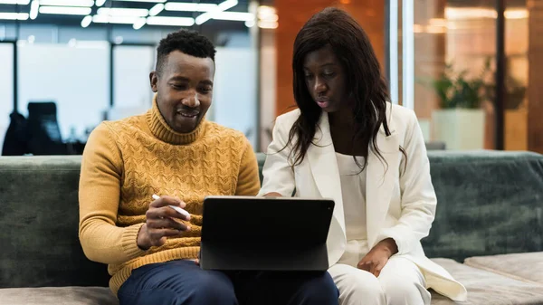 Black man and woman in an office working using a tablet while sitting on a sofa