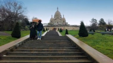 PARIS, FRANCE - JANUARY, 2022: View of The Basilica of Sacre Coeur from the staircase in front of it, walking people, bare trees, cloudy sky