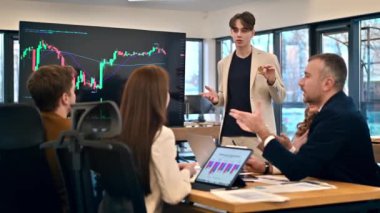 Young worker leading business meeting in an office, discussing the topic of cryptocurrencies with other workers using a big display with currency rate and physical coin. Slow motion