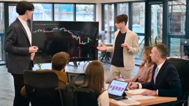 Two young workers leading business meeting in an office, discussing the topic of cryptocurrencies with other workers using a big display with currency rate and physical coins. Slow motion