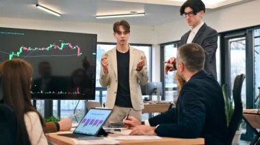 Two young workers leading business meeting in an office, discussing the topic of cryptocurrencies with other workers using a big display with currency rate and physical coins. Slow motion