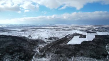 Aerial drone view of a nature in Moldova in winter. Fields and hills covered with snow, forest plantings with bare trees, village in the distance