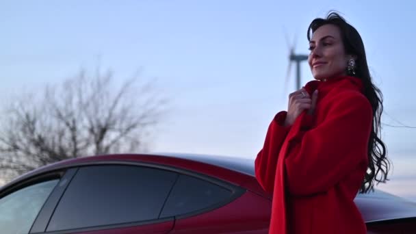 日没時に赤い車と風力タービンの近くにポーズをとる幸せな女性 — ストック動画