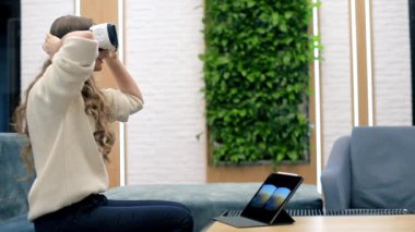 Sarışın, beyaz bir kadın VR gözlük takıyor ve ofiste sanal gerçekliği keşfetmeye başlıyor. Tablet masanın üzerinde, resim kulaklıkta görünür.