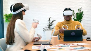 Business conference in VR in an office. A black man and caucasian woman using VR glasses and controllers, papers and gadgets on the table