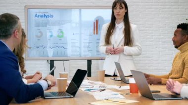 Ofiste bir iş toplantısında kadın takım lideri. İşçilerle iş meselelerini tartışıyor. Çizelgeli büyük bir sergi, kağıtlı masa ve aletler kullanıyor.