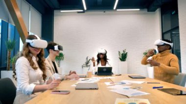 Business conference in VR in an office. Multiracial group of people using VR glasses and controllers, papers and gadgets on the table