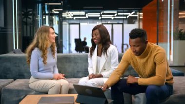 Siyahi bir adam bir ofiste kanepede oturup tablet kullanırken, beyaz ve siyah kadınlar iş konuşuyorlar.