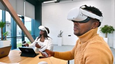 VR 'da bir ofiste iş konferansı. Siyah bir adam ve kadın VR gözlük ve kumanda kullanıyor. Masada aletler var. Yavaş çekim