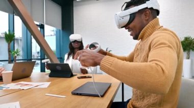 VR 'da bir ofiste iş konferansı. Siyah bir adam ve kadın VR gözlük ve kumanda kullanıyor. Masada aletler var. Yavaş çekim