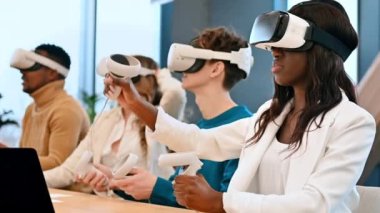 VR 'da bir ofiste iş konferansı. Sanal gerçeklik gözlüğü ve kumanda kullanan çok ırklı bir grup insan, masada aletler. Yavaş çekim