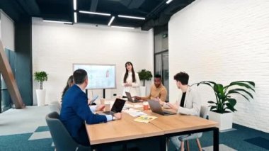 Ofisteki bir iş toplantısında kadın takım lideri, masa üzerinde tablolar, kağıtlar ve aletler olan büyük bir vitrin kullanarak işçilerle iş ilişkilerini tartışıyor.