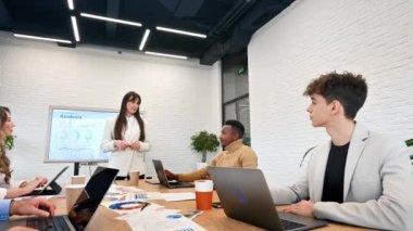 Ofisteki bir iş toplantısında kadın takım lideri, masa üzerinde tablolar, kağıtlar ve aletler olan büyük bir vitrin kullanarak işçilerle iş ilişkilerini tartışıyor.