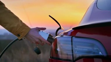 Gün batımında park halindeki kırmızı elektrikli bir arabanın şarj kablosunu çeken adamın yakın görüntüsü.