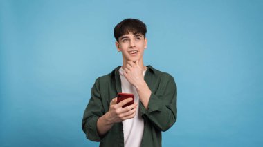 Telefonunu mavi arka planda tutan genç bir adam.
