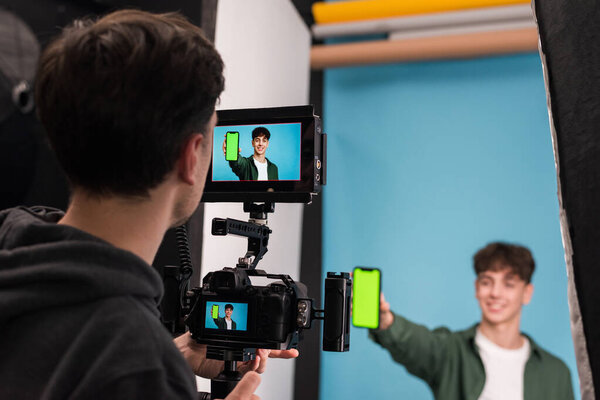 View of a professional photographer shooting a young man showing smartphone with green screen, blue background