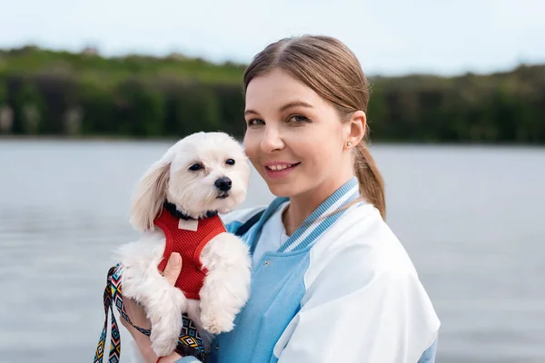 View of a blonde woman holding her maltese dog with white fur and looking in the camera, lake on the background