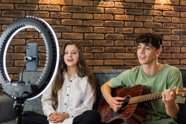 Genç bir adam ve kadın gitar çalıp şarkı söylüyor. Stüdyoda yüzüklü bir ışıkla akıllı telefondan kendilerini vuruyorlar.