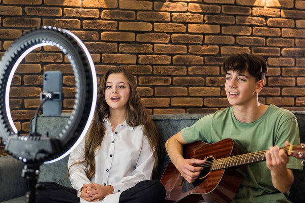 Young smiling man and woman playing on a guitar and singing, shooting themselves on smartphone on a tripod with ring light in a studio