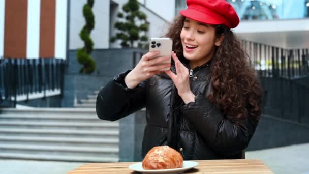 スマートフォンでクロワッサンを撮影した赤い帽子の若い女性ブロガーの影響力 スローモーション — ストック動画