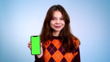 Gülümseyen ve yeşil ekranlı telefonunu gri arka planda kameraya gösteren genç bir kadın.