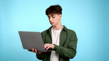 Genç adam dizüstü bilgisayarını kullanıyor ve mavi arka planda gülümsüyor.