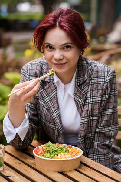 stock image Portrait of a smiling young woman eating poke bowl with vegetables and fruits, holding a bean with chopsticks. Park with a lot of greenery and illumination