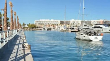 Alicante Limanı 'nda yüzen bir tekne ve palmiye ağaçlarıyla sokakta yürüyen insanlar görülüyor.