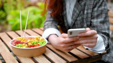 Telefonunu kullanan bir kadının yakın görüntüsü, masada sebze, meyve ve çubuk dolu bir kase. Bir sürü yeşillikle park et. Yavaş çekim