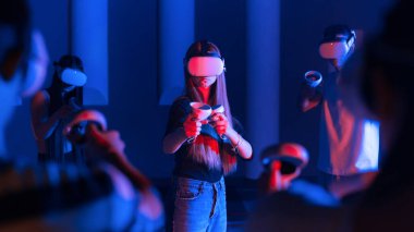 Sanal gerçeklik VR ekipmanlarını bir arenada kullanan bir grup genç arkadaş. Merkezde bir kız ve arka planda diğer oyun arkadaşları. Neon ışıkları