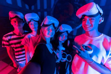 Bir grup genç arkadaş arenada sanal gerçeklik VR ekipmanı kullanırken selfie çekiyorlar. Neon ışıkları
