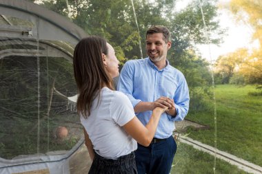 Şeffaf kabarcık çadırında sarılan bir çift. Çevredeki yemyeşil orman ve doğa