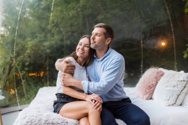 Yatakta otururken şeffaf kabarcık çadırında kucaklaşan bir çift. Arka planda yemyeşil orman